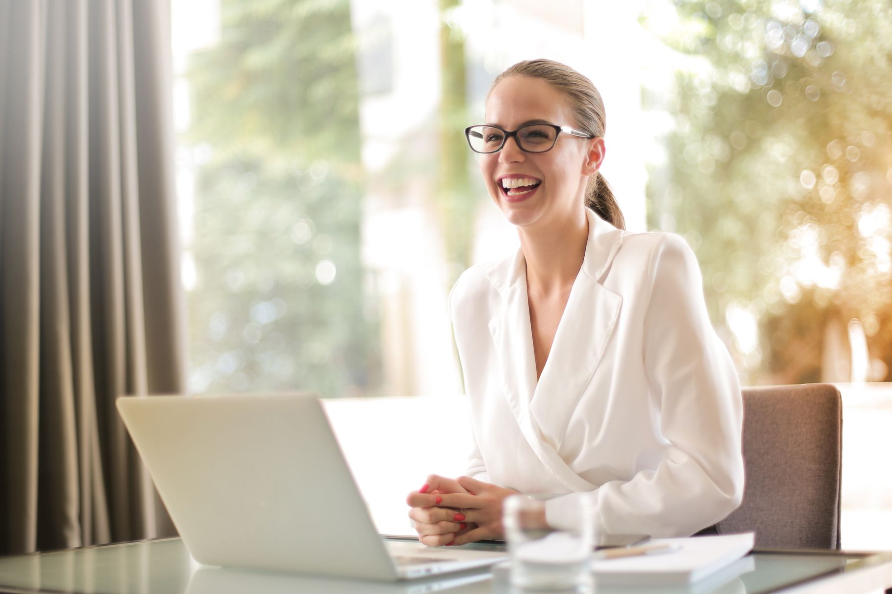 Lachende vrouw met bril achter een laptop bij een raam